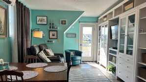 A dining area with a wooden table and chairs, a blue armchair, and a teal wall with framed artwork and a door leading outside.