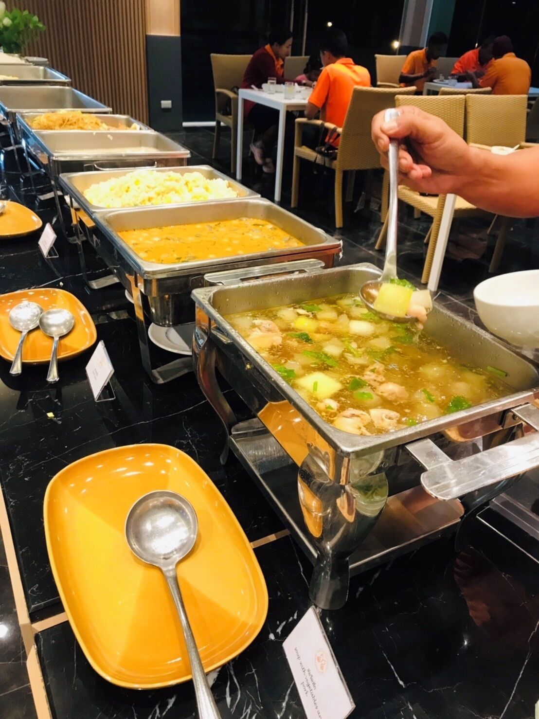 Montaje de bufé con diversas opciones de comida, incluyendo sopa de verduras y carne, y una persona servándose.