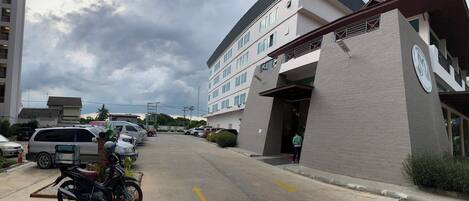 A parking lot with several cars and a motorcycle, a building with a clock on the side, and a person standing near the entrance.