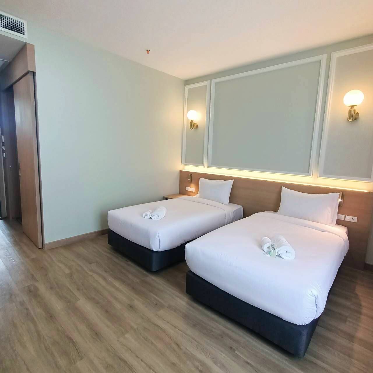 A hotel room with two beds, each with white linens and towels, a wooden floor, and wall-mounted lighting.