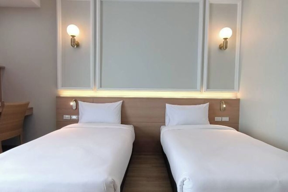 A hotel room with two single beds, a wooden headboard, bedside tables, and wall-mounted light fixtures.