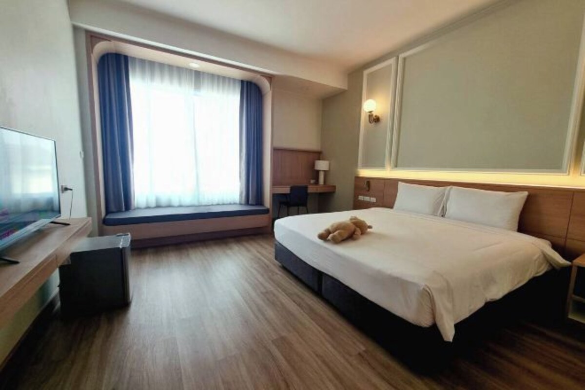 A hotel room with a bed, a TV, a window with curtains, and a desk with a lamp.