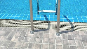 Una piscina con escaleras de metal, rodeada por un patio de baldosas y un edificio con un mural geométrico.