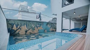 Una piscina con un mural de camuflaje, un área de descanso con sillas azules y vista a un edificio al fondo.