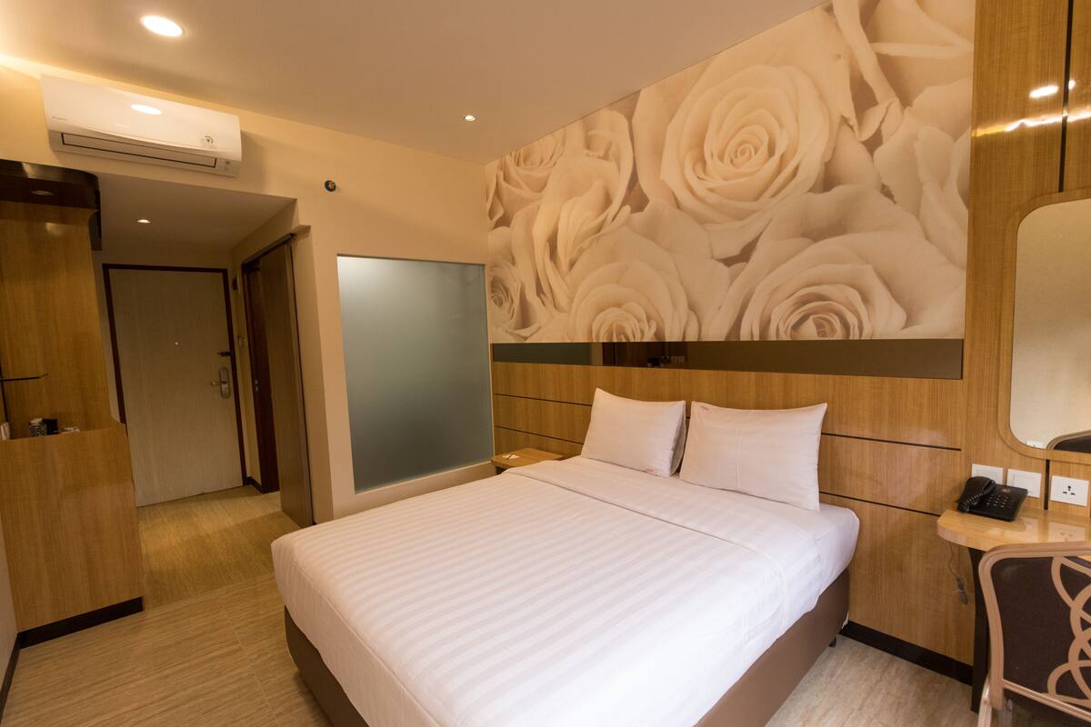 A hotel room with a large bed, a wooden headboard, a wall with a floral wallpaper, a bedside table with a phone, and an air conditioning unit on the ceiling.