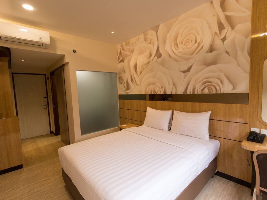 A hotel room with a large bed, a wooden headboard, a wall with a floral wallpaper, a bedside table with a phone, and an air conditioning unit on the ceiling.