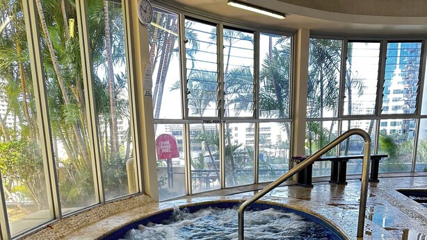 A hot tub with a metal ladder, surrounded by a tiled floor and large windows with a view of palm trees and buildings. - Chevron Renaissance-Resort Style Living managed by Gold Coast Premium (Surfers Paradise)