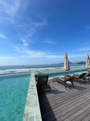 Ein Infinity-Pool mit Blick auf das Meer, eine Holzterrasse und Liegestühle mit Sonnenschirmen.