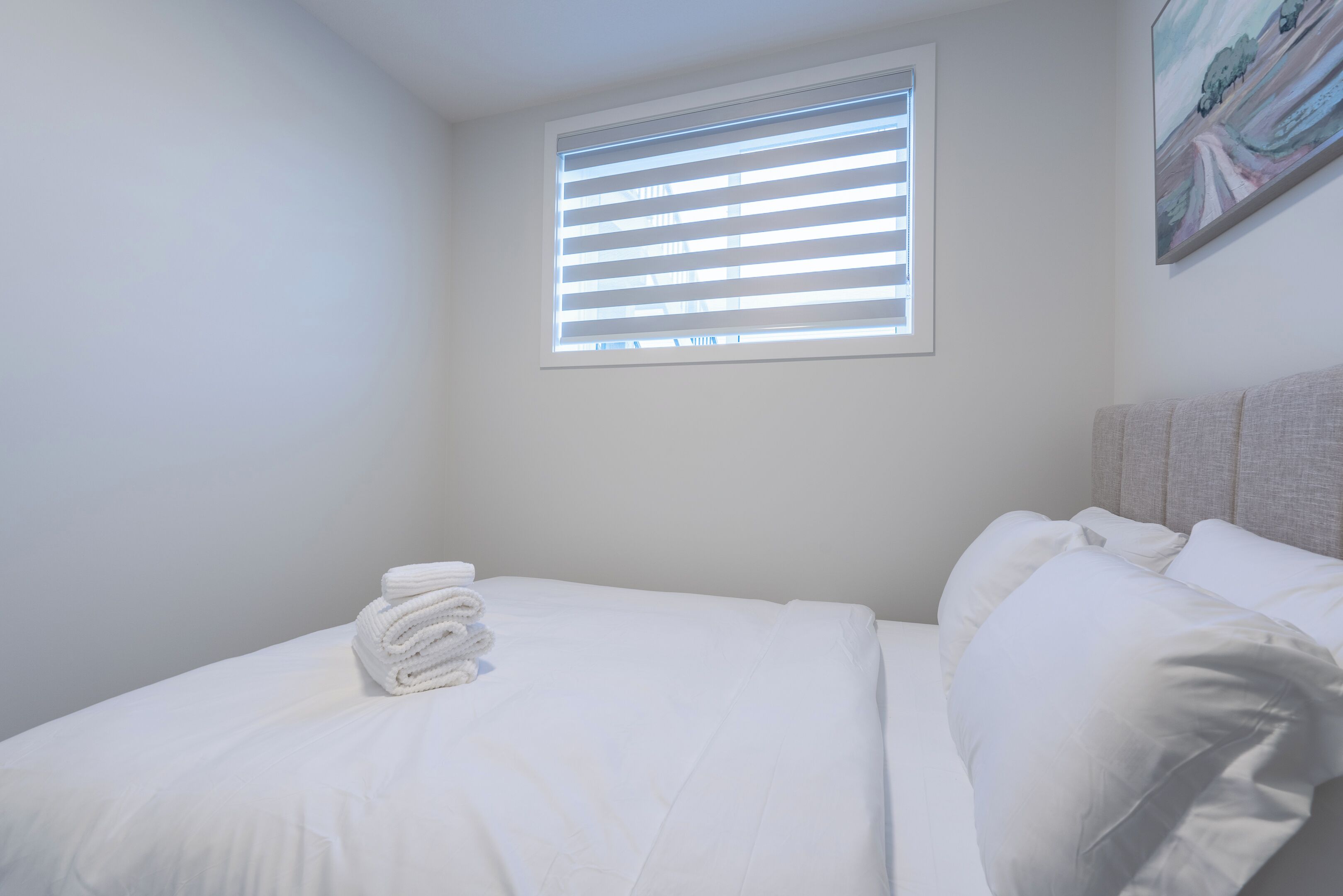 A neatly made bed with white linens and a rolled towel, a window with blinds, and a painting on the wall.