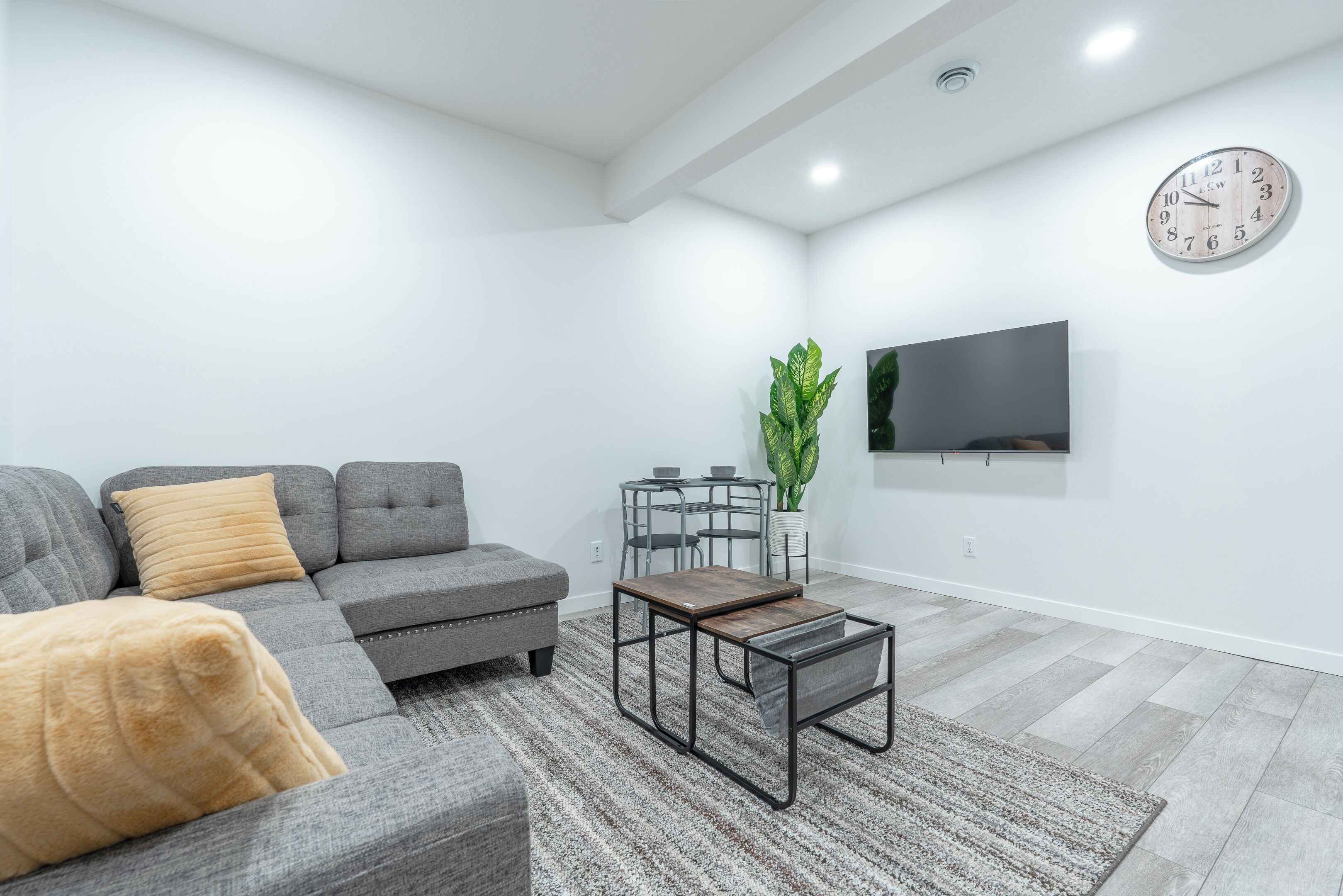 Ein modernes Wohnzimmer mit einem grauen Ecksofa, einem kleinen Couchtisch, einem an der Wand befestigten Fernseher und einer Zimmerpflanze.