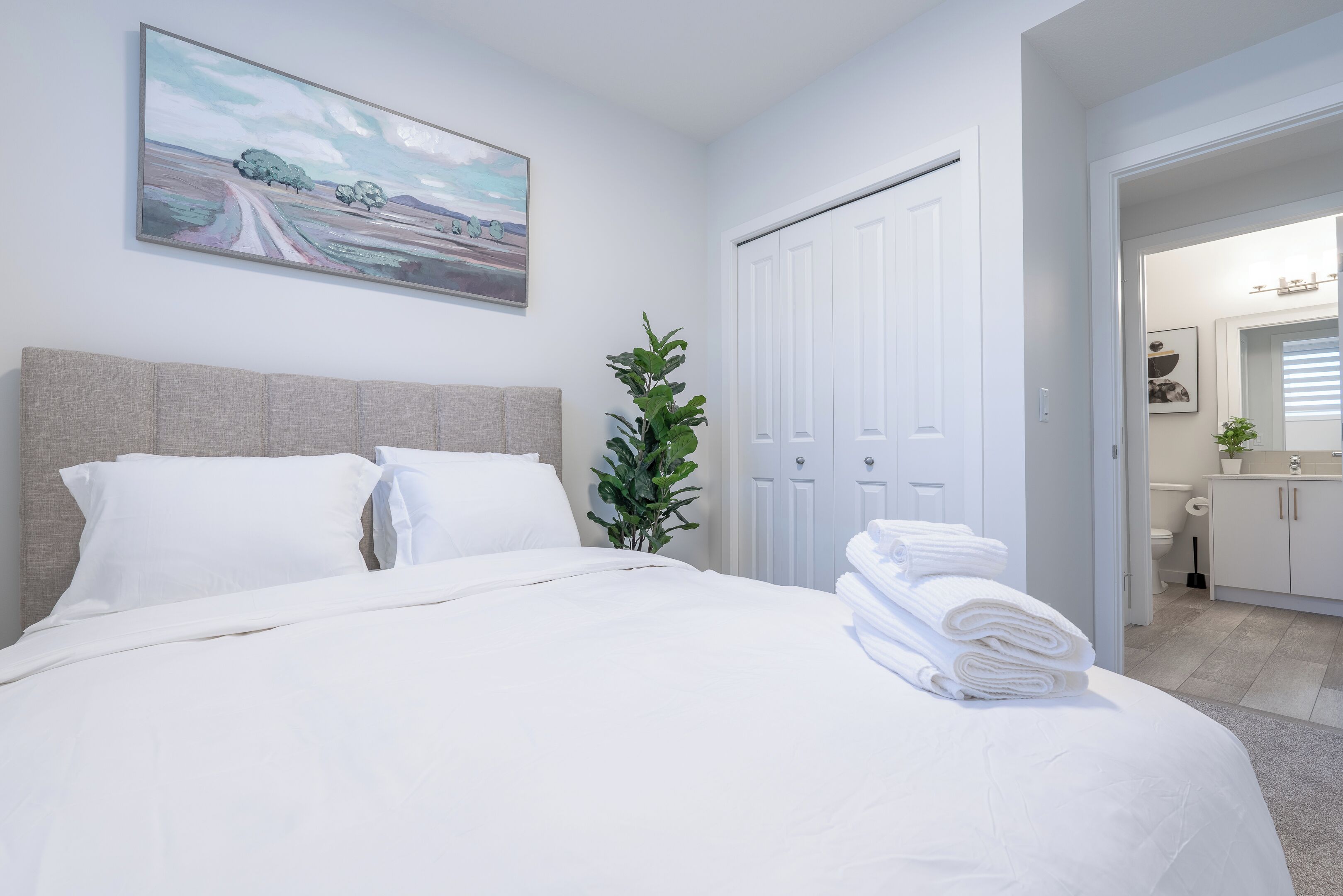 Una cama bien tendida con sábanas blancas, un cuadro grande de un paisaje en la pared y una puerta que da a un baño.