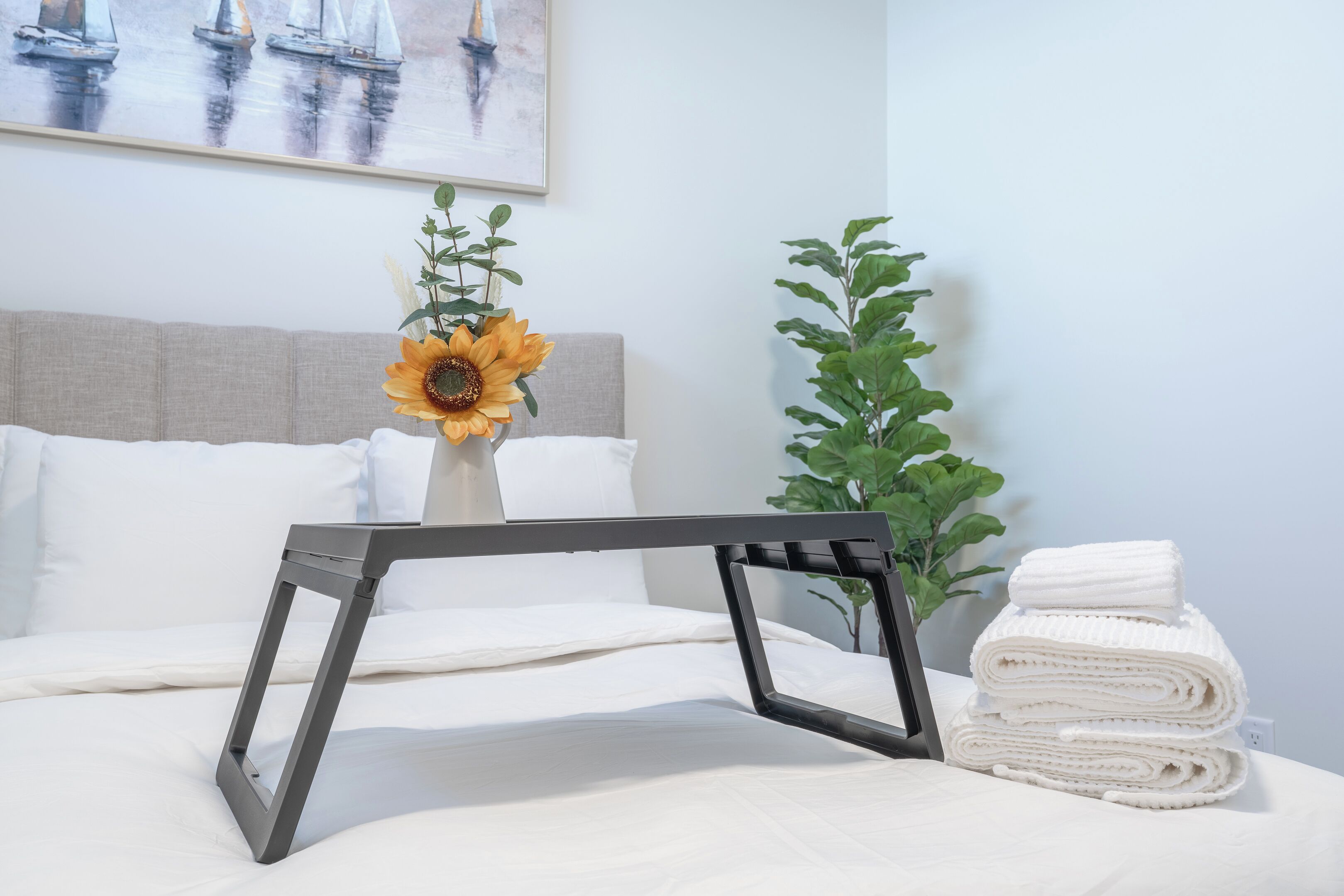 Un lit bien fait, avec du linge de lit blanc, un cadre en métal noir, un vase contenant un tournesol et une pile de serviettes blanches.