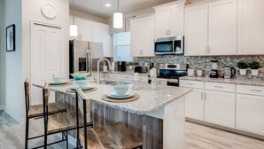 A modern kitchen with white cabinets, granite countertops, a central island with bar stools, a microwave, and a refrigerator. - Luxury 5BR Home, Private Pool, Prime Location (Kissimmee)