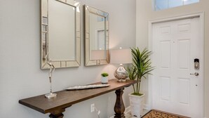 A hallway with a wooden console table, two wall mirrors, a potted plant, a lamp, and a white door with a doormat. - Luxury 5BR Home, Private Pool, Prime Location (Kissimmee)