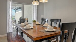 Un coin repas avec une table en bois dressée pour quatre personnes, deux lampes suspendues et une vue sur un balcon à travers une porte vitrée.
