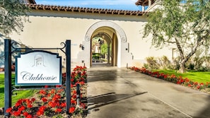 Entrada do Clubhouse com um letreiro, uma porta em arco e um caminho bordeado por flores vermelhas.