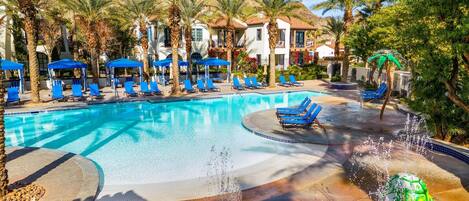 A swimming pool with blue lounge chairs and palm trees, surrounded by buildings with a Mediterranean architectural style.