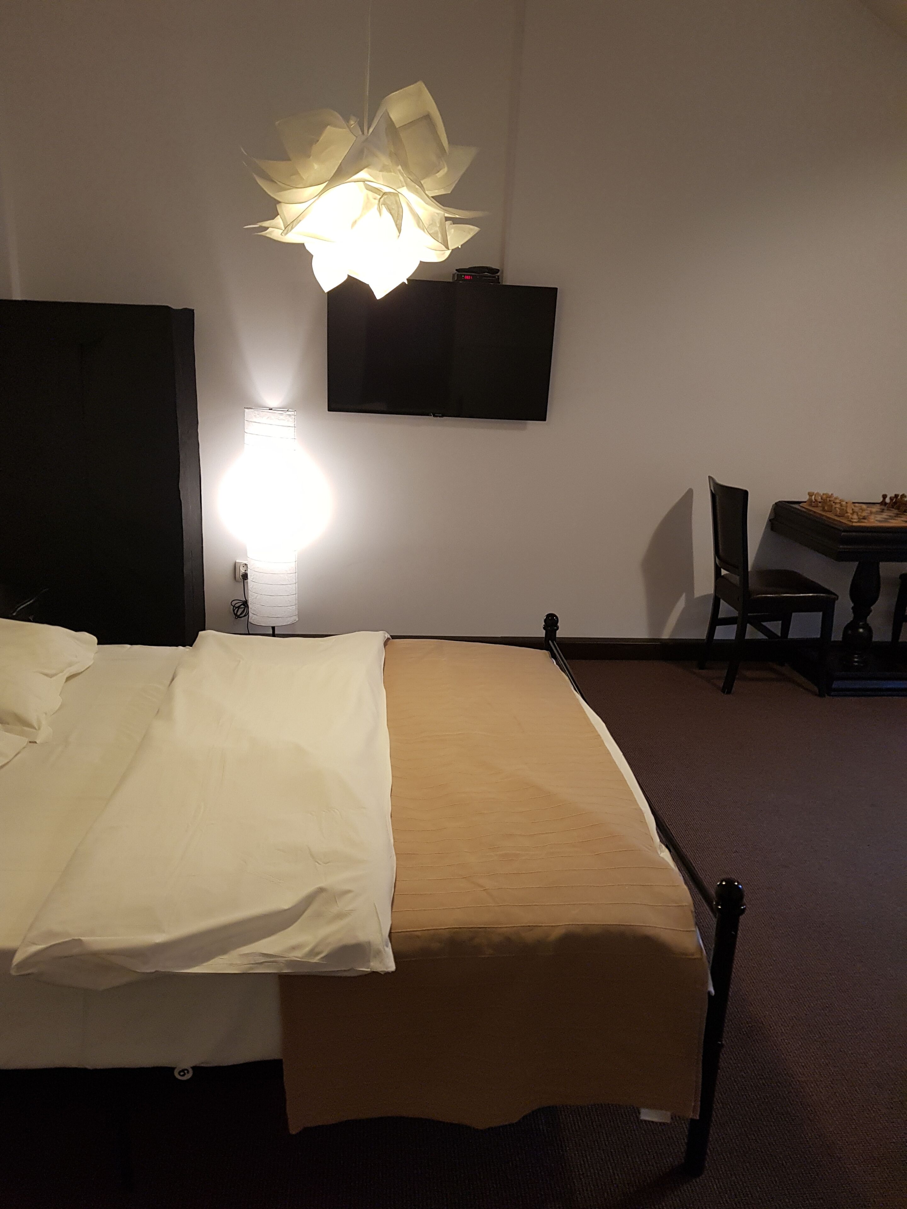 A hotel room with a bed, a desk with a chessboard, a television, and a unique ceiling light fixture.