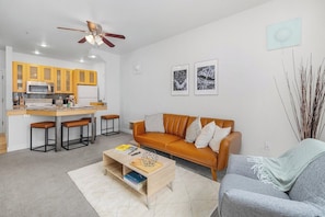 A modern living room with a brown leather sofa, a grey armchair, a wooden coffee table, and a kitchen area with yellow cabinets and a microwave.