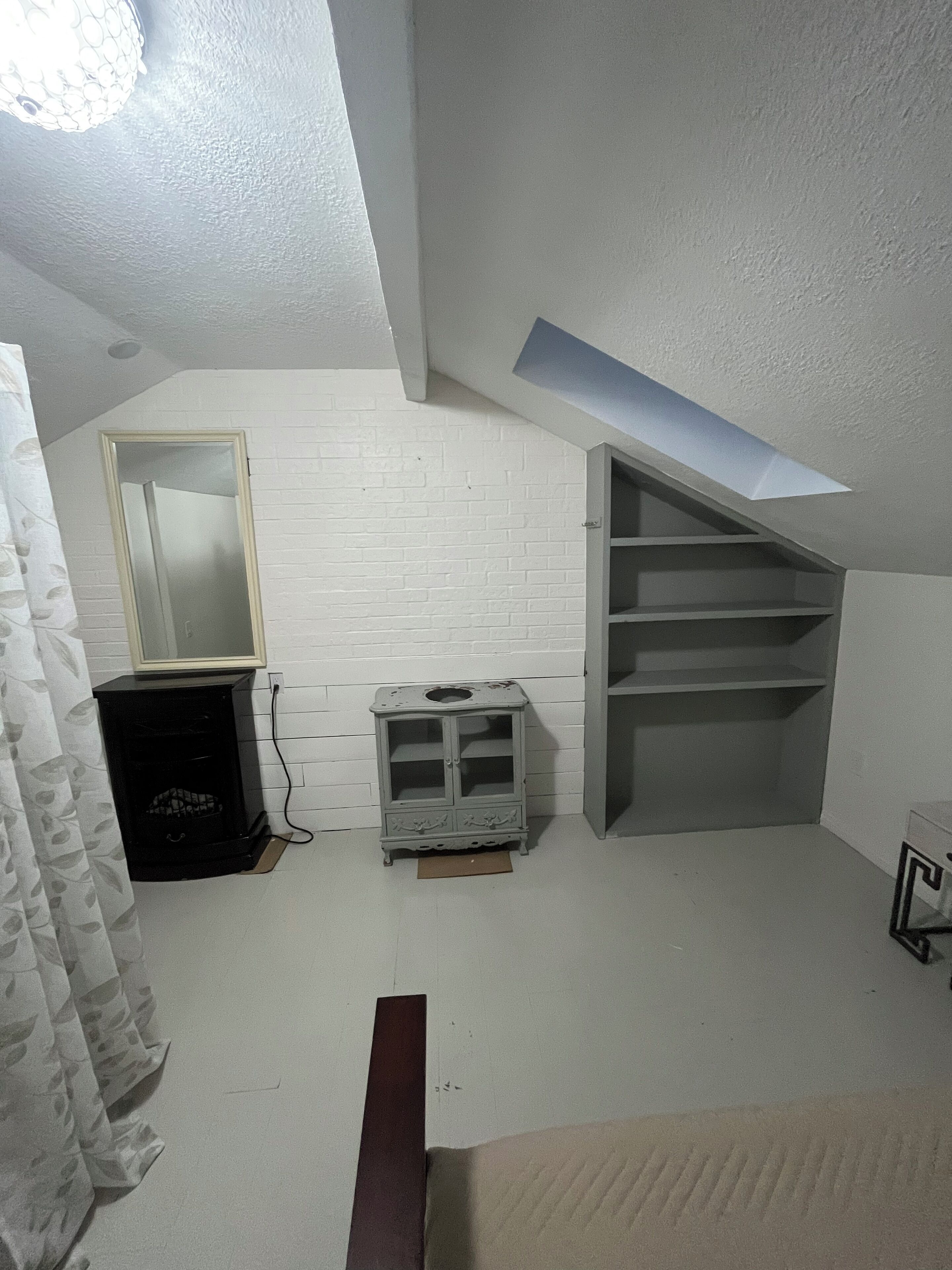 Una habitación con techo inclinado, pared de ladrillo blanco, un mueble pequeño, un espejo y una estantería.