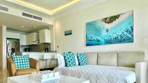 A modern living room with a sofa, coffee table, and a painting of a beach scene. - Luxurious, Stunning Bay View Condo, King Bed, Spacious Terrace, Pool, Gym. (Samaná)