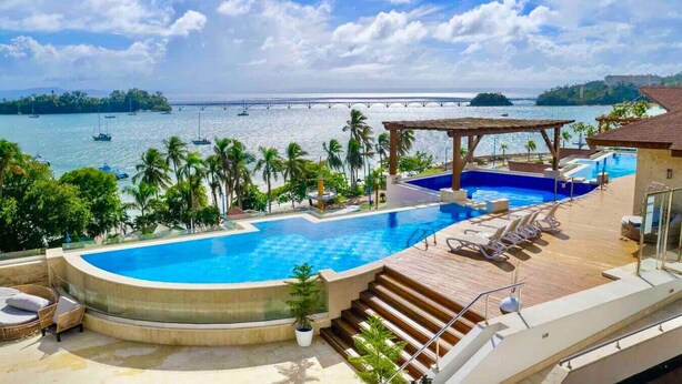 A pool area with a view of the sea, a covered seating area, and a wooden deck. - Luxurious, Stunning Bay View Condo, King Bed, Spacious Terrace, Pool, Gym. (Samaná)
