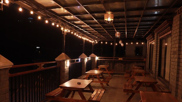 An outdoor patio with wooden tables and benches, string lights, and brick walls. - Hotel De Purang (Muktinath)