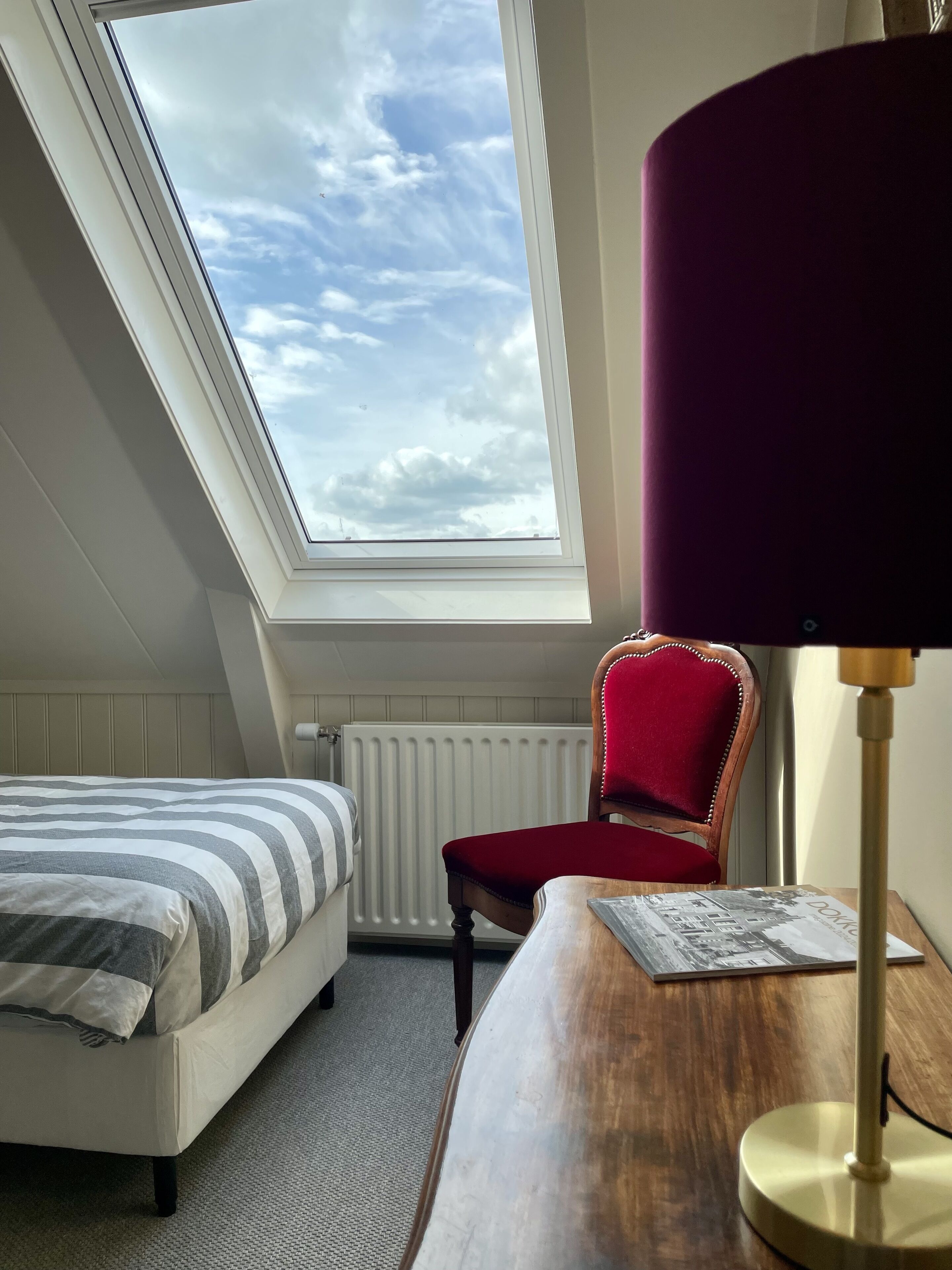 A bedroom with a skylight, a bed with striped bedding, a red chair, and a wooden desk with a lamp.