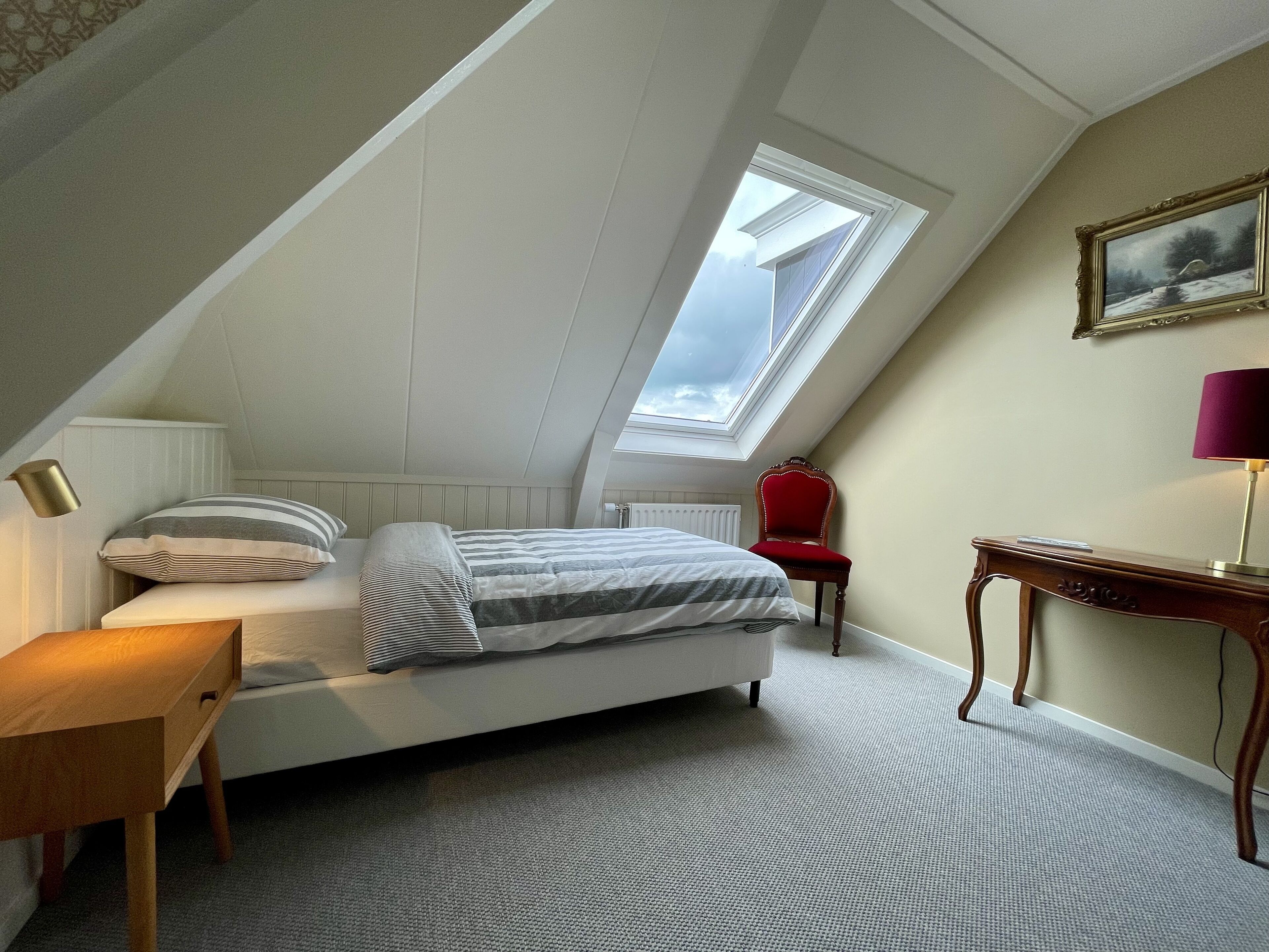 Une chambre avec un plafond en pente, un puits de lumière, un lit avec une literie à rayures, une chaise rouge, un bureau en bois et un tableau encadré accroché au mur.