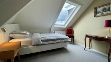 Ein Schlafzimmer mit schrägem Dach, einem Oberlicht, einem Bett mit gestreifter Bettwäsche, einem roten Sessel, einem Holztisch und einem gerahmten Bild an der Wand.