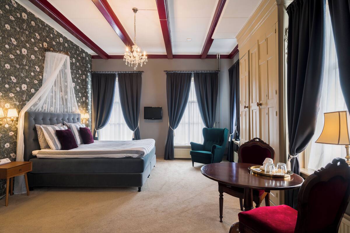 Habitación de hotel con una cama grande, una zona de estar con una mesa y sillas, una araña y un televisor.