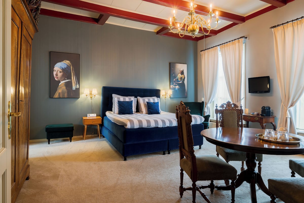 Habitación de hotel con una cama grande, una mesa de comedor, una araña y obras de arte en las paredes.
