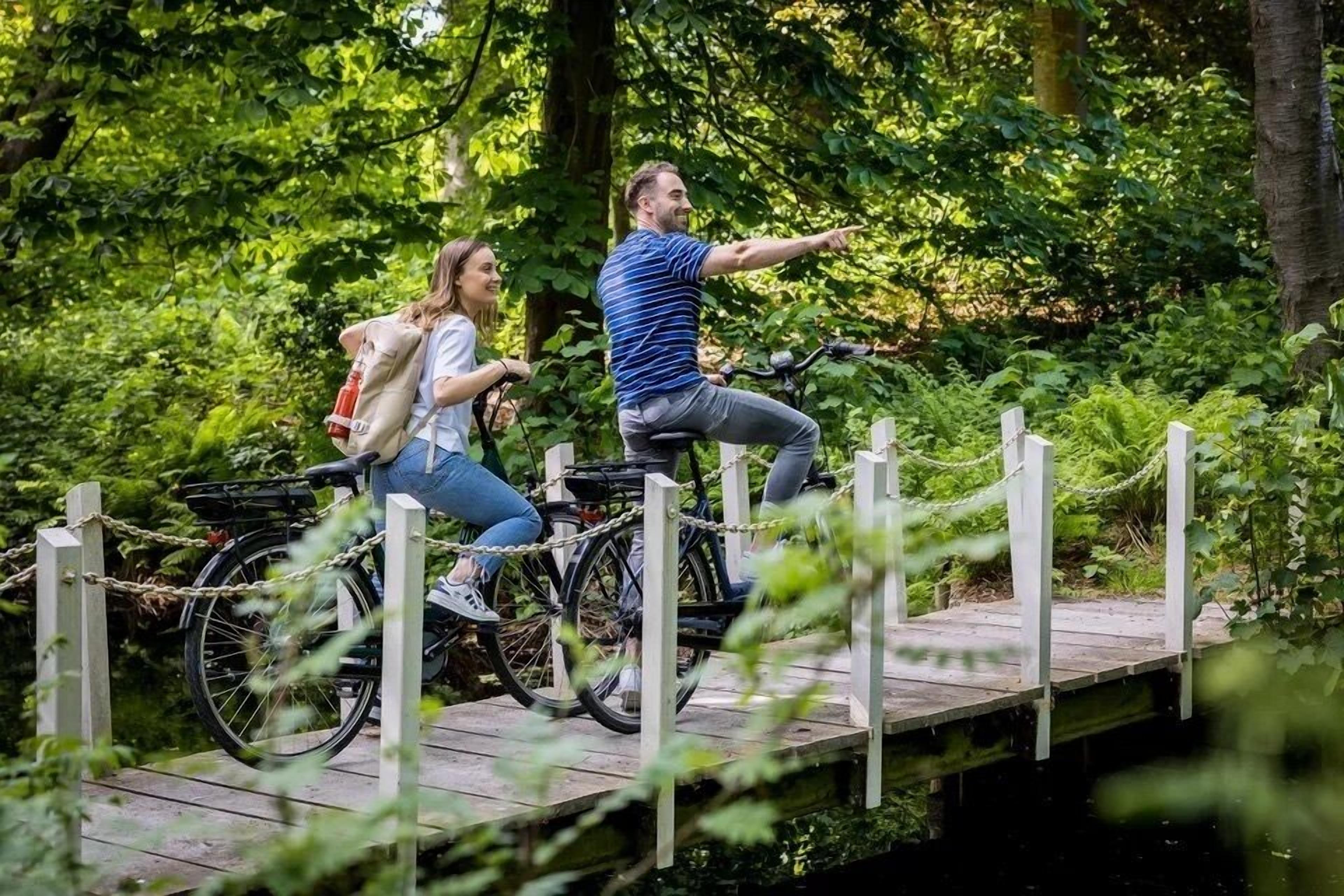 Zwei Personen fahren auf Fahrrädern über eine Holzbrücke, umgeben von viel Grün.
