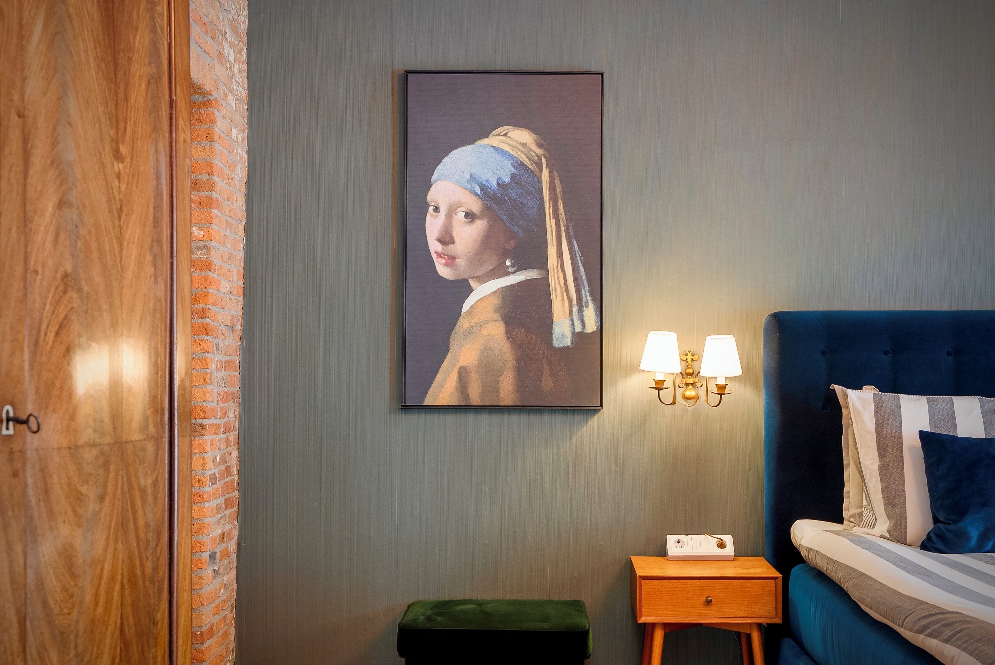A bedroom with a bed, a nightstand, a green bench, a wall-mounted painting, and a wooden door.