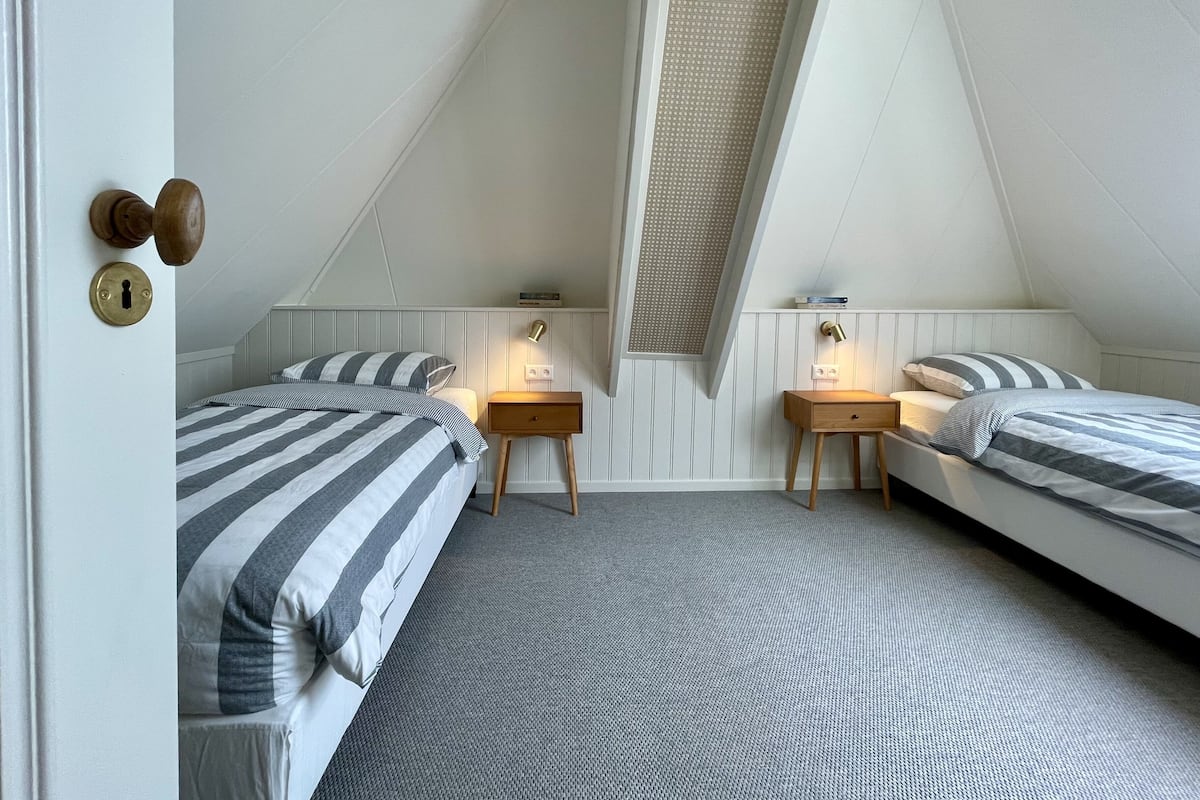 Un dormitorio ático pequeño y triangular con dos camas, cada una con ropa de cama a rayas, dos mesitas de noche con lámparas y un techo inclinado con tragaluz.