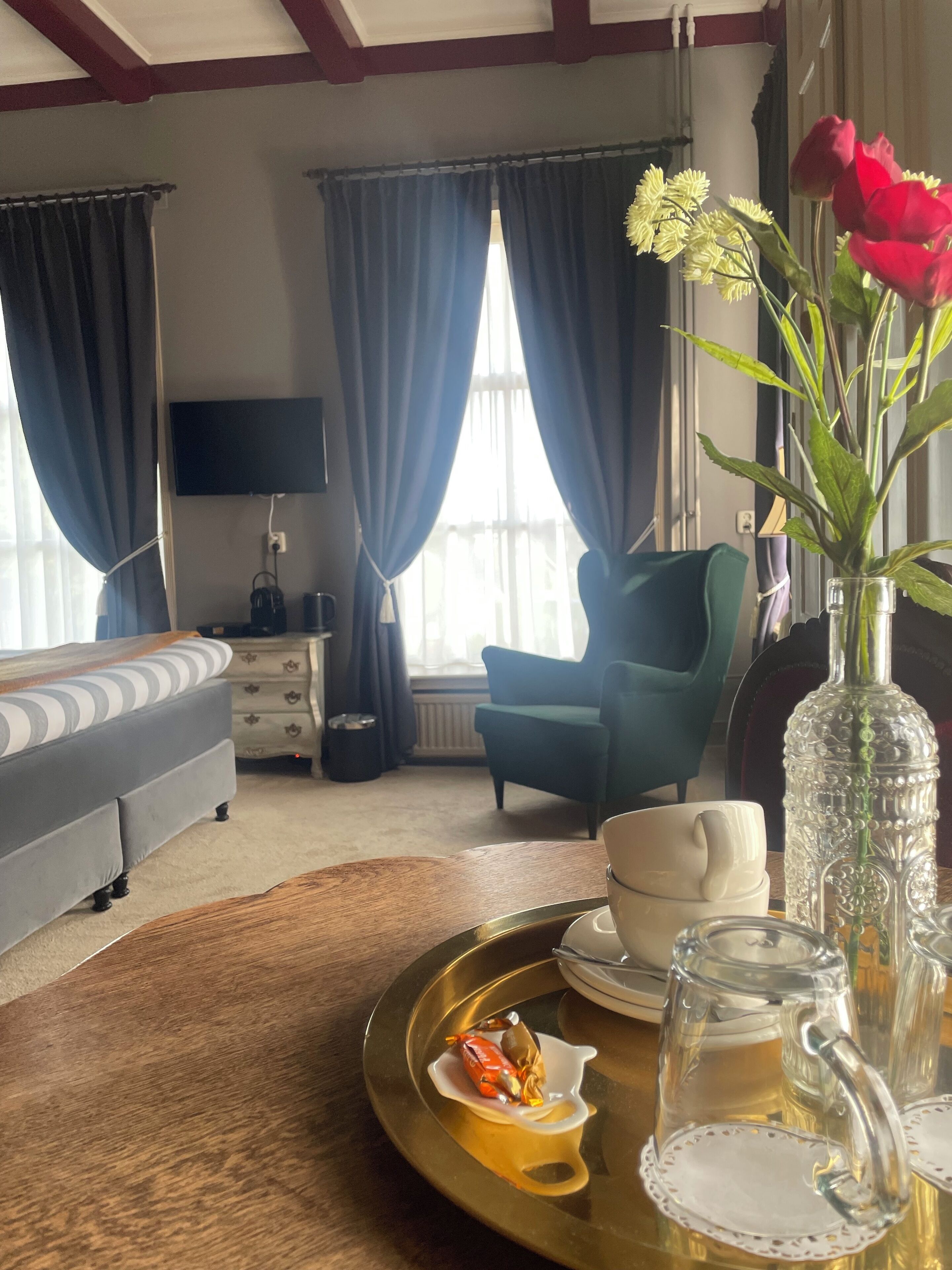 A hotel room with a bed, a TV, a dining table with a tray, and a vase with flowers.
