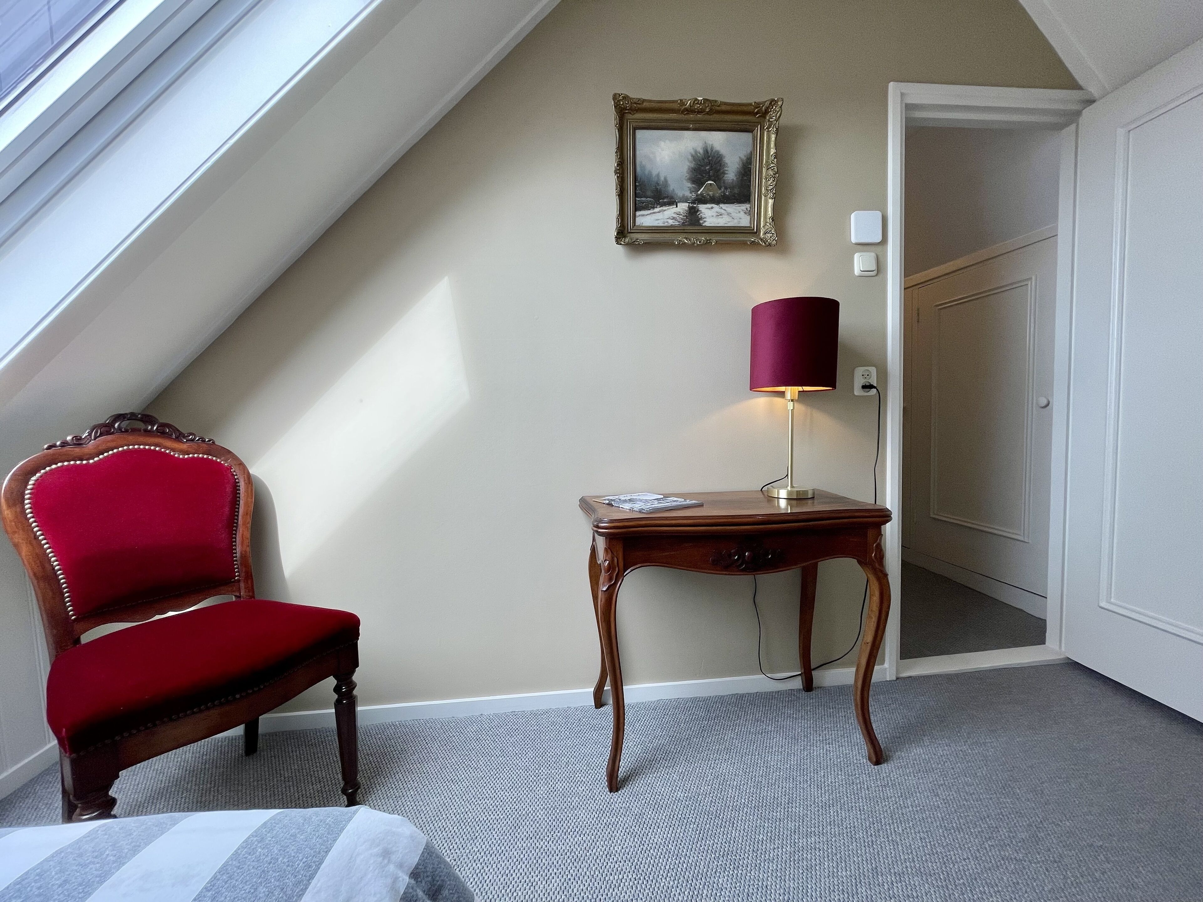 A room with a sloped ceiling, a red upholstered chair, a small wooden table with a lamp, and a framed picture on the wall.