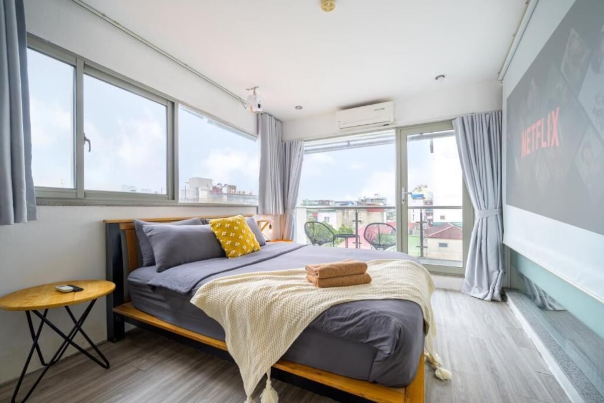 A modern bedroom with a large bed, a small table, and a view of the city through large windows.