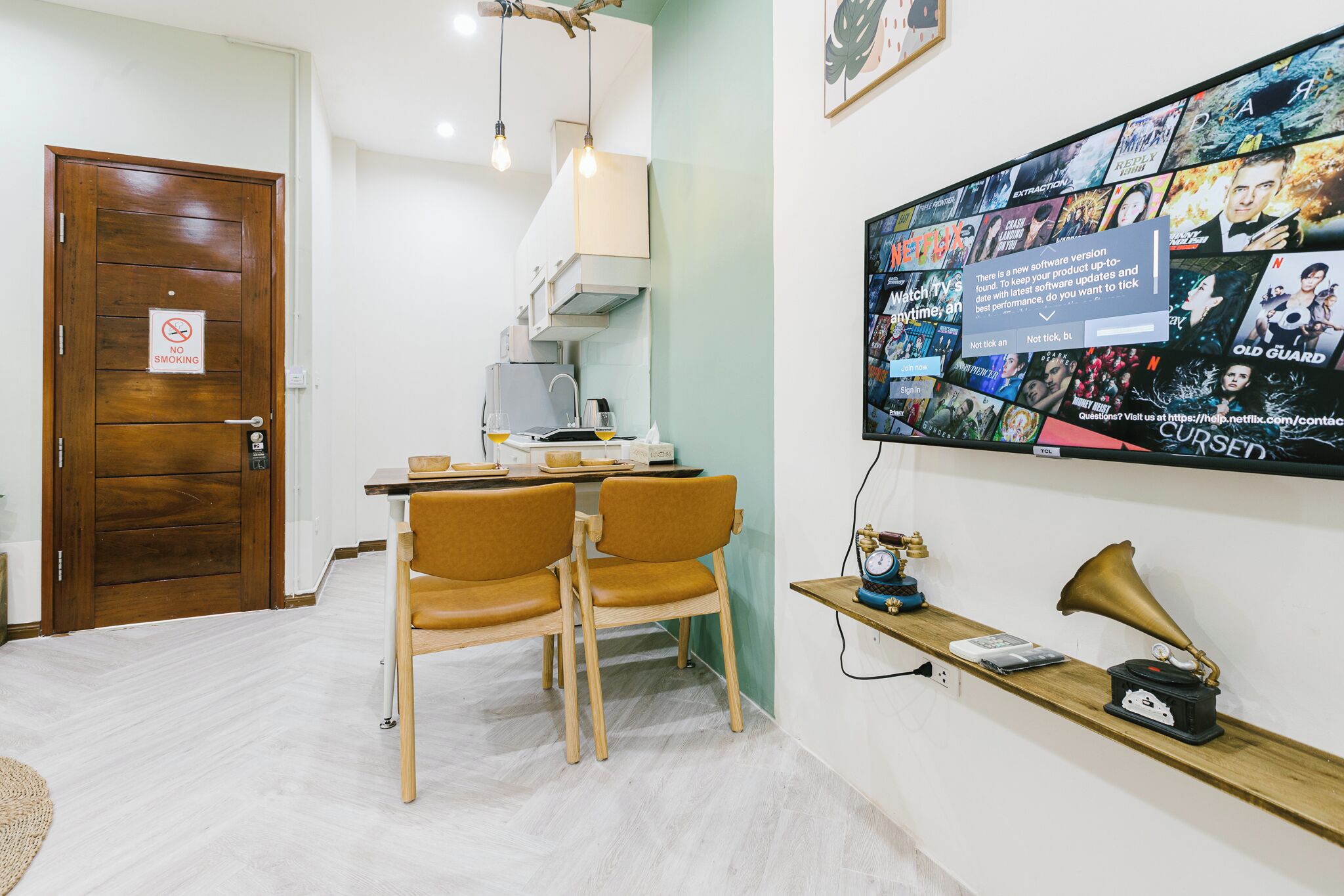 A room with a wooden door, a dining table with chairs, a wall-mounted TV displaying a Netflix interface, and a shelf with a gramophone and other items.