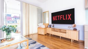 Un salon moderne avec une grande télévision affichant le logo « NETFLIX », un meuble en bois, une table basse en verre et une vue sur l’extérieur grâce à une porte coulissante.
