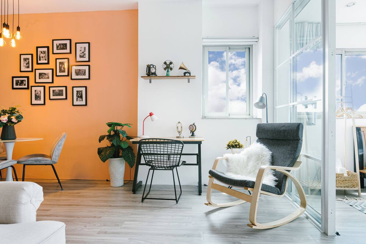 A modern living room with a dining area, a rocking chair, and a large window with a view of the sky.