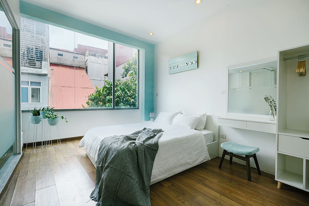 A modern bedroom with a large window, a bed, a small stool, and a white shelf unit.