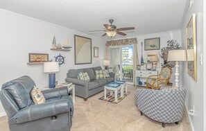 Relax in a stylish living room with a leather armchair, sofa, and ottoman, surrounded by nautical-themed decor and a large window with an outdoor view.