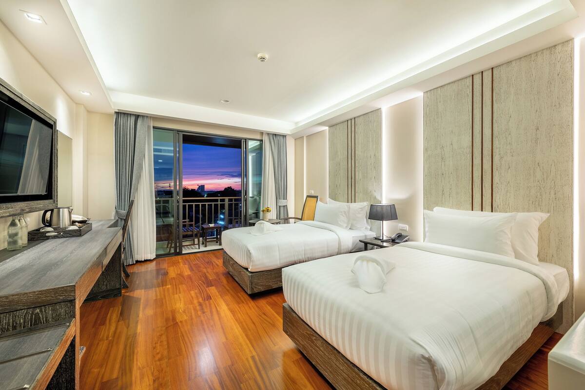 A hotel room with two beds, a TV, a desk, and a balcony with a city view.