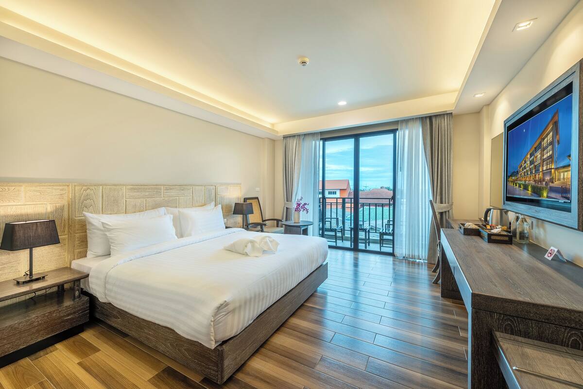 A hotel room with a large bed, a desk, a TV, and a balcony with a view of buildings.
