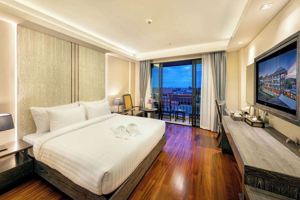 A hotel room with a large bed, a TV, a balcony with a city view, and a wooden floor.