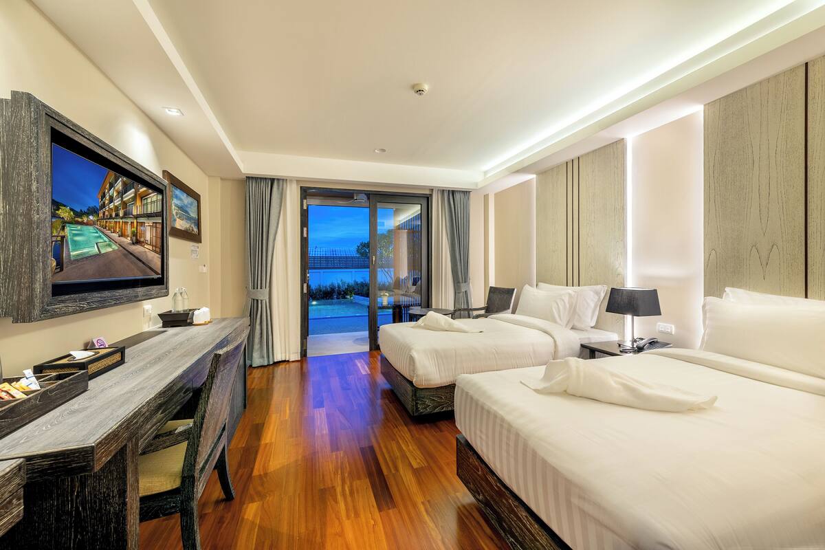 A hotel room with a wooden desk, two beds, a TV, and a view of a pool and building.