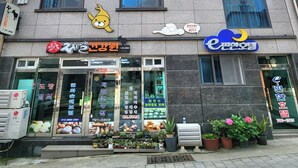 Un edificio de varios pisos con diferentes locales comerciales, incluyendo un restaurante visible y una tienda con un letrero de un personaje de dibujos animados.