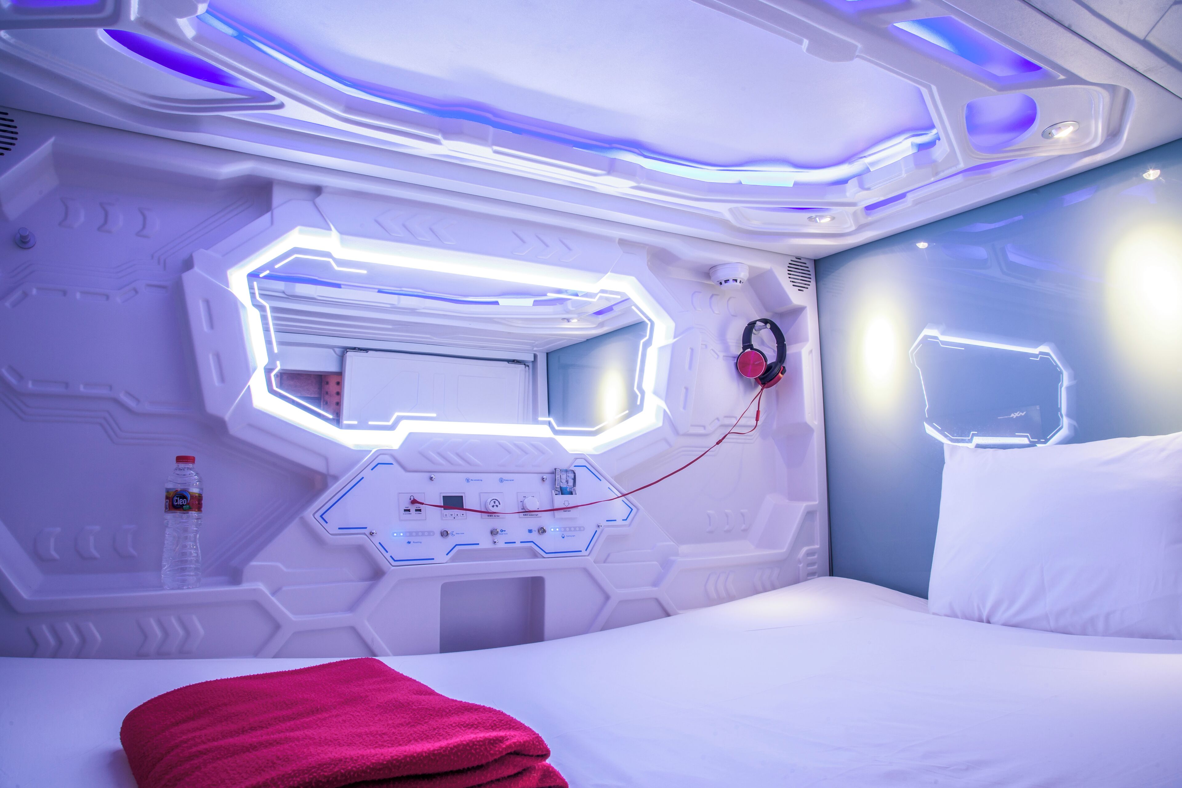 Habitación de hotel moderna con una cama blanca, una manta roja, una botella de agua y un par de auriculares en la mesita de noche.
