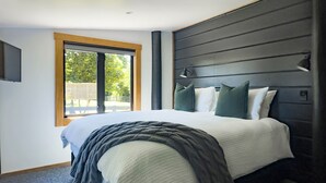 A modern bedroom with a large bed, a window with a view of greenery, and a dark headboard.